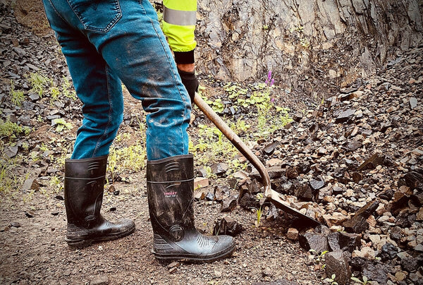 Person wearing blue jeans and black boots with a yellow vest, standing on rocky terrain and holding a shovel.