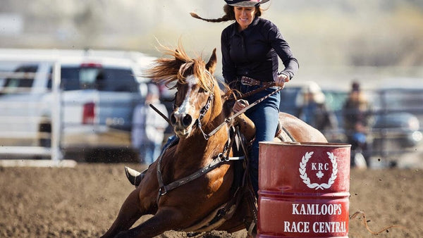 woman in western clothes and black cowgirl hat riding brown horse around a barrel in a dirt arena