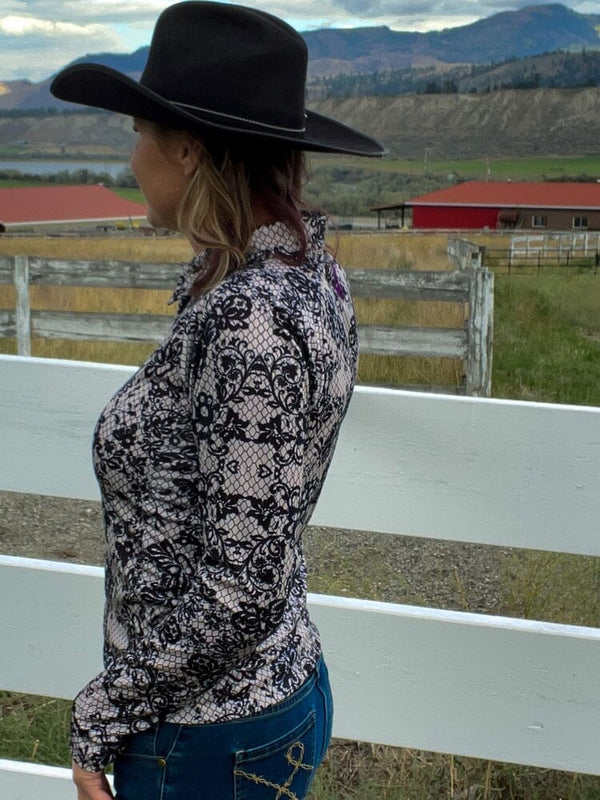 woman outside turned to the side, standing by white wood fence, wearing black and white lace pattern shirt and black western hat