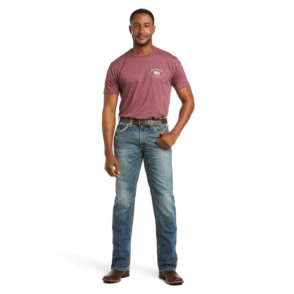full body view of man wearing medium wash distressed denim jeans and a maroon short sleeve t-shirt, with brown belt and boots