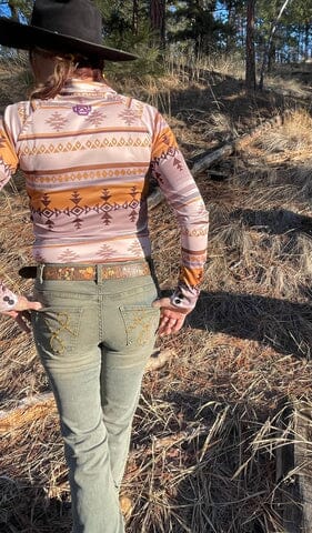 back of woman with thumbs in back pockets, wearing cream and brown patterned long sleeve button up shirt, light grey pants, and black cowgirl hat