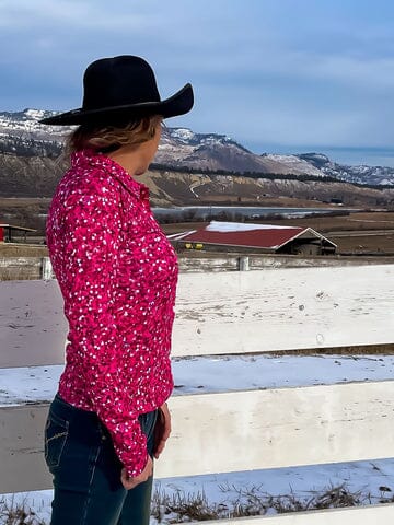 woman outside standing by white wood fence, wearing pink and white glitter pattern shirt and black western hat