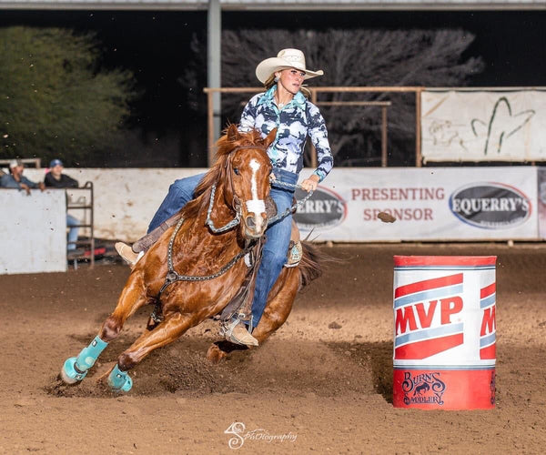 woman in western clothes and cowgirl hat riding brown horse around a barrel in a dirt arena