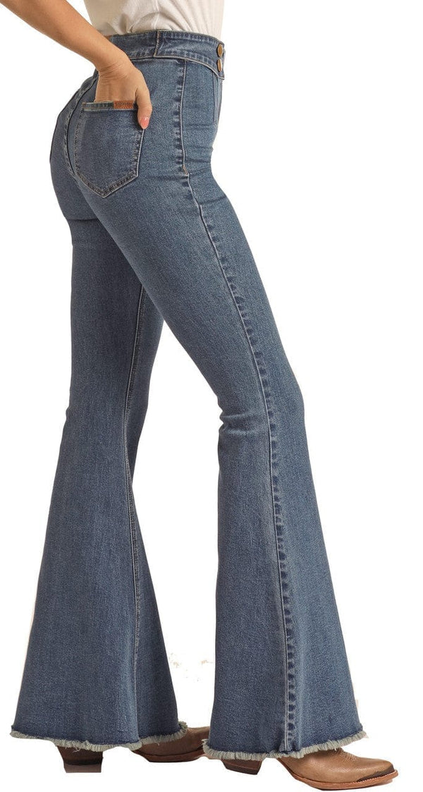 side view of woman wearing medium blue bell bottom jeans, with button details on waistband and tan point toe boots, with hand in back pocket
