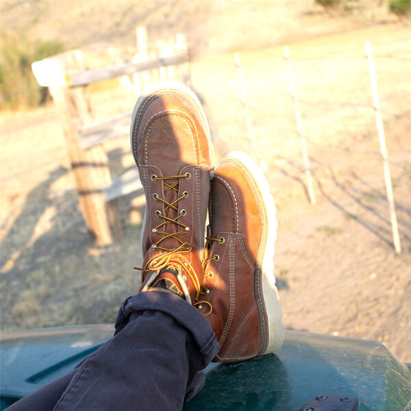 crossed feet wearing brown/red mid-rise boot with yellow and brown laces and white sole