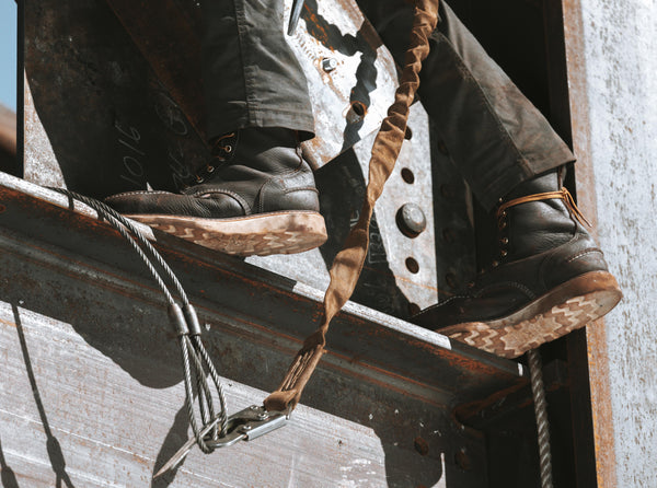 person standing on edge of metal beam while wearing brown high top moccasin style boots with worn white soles exposed