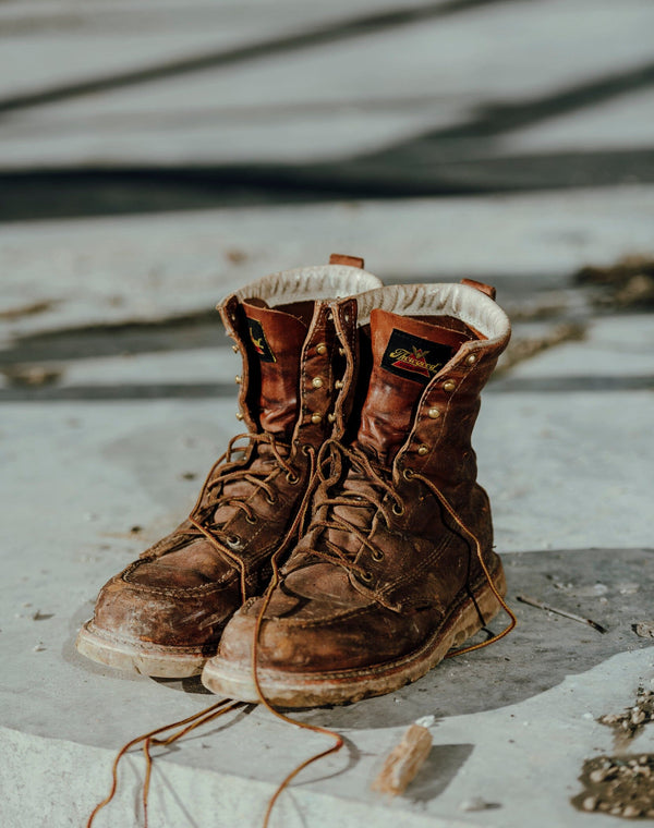 pair of worn-looking mens brown logger boot on concrete surface, with white interior and muddy sole. Brown/gold laces half unlaced.