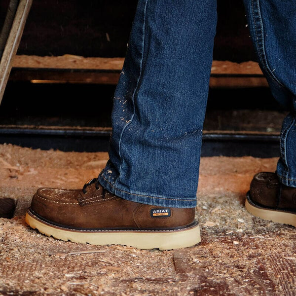 Person waring brown Ariat work boots with blue jeans on a wooden dirty floor.