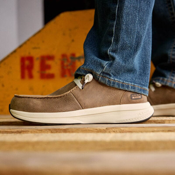 Brown shoe with white sole worn by a person in blue jeans, standing on a wooden surface.