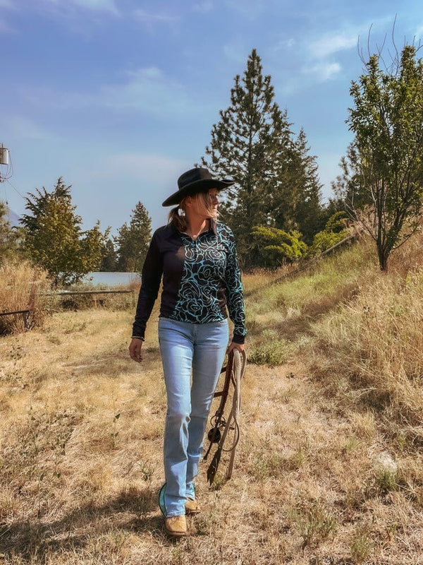 woman walking outside holding a harness wearing light blue jeans, black western hat, and black and turquoise long sleeve zip up shirt with rose print