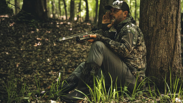 Hunter wearing camouflage and brown and camouflage boots sitting against a tree in a forest with a rifle