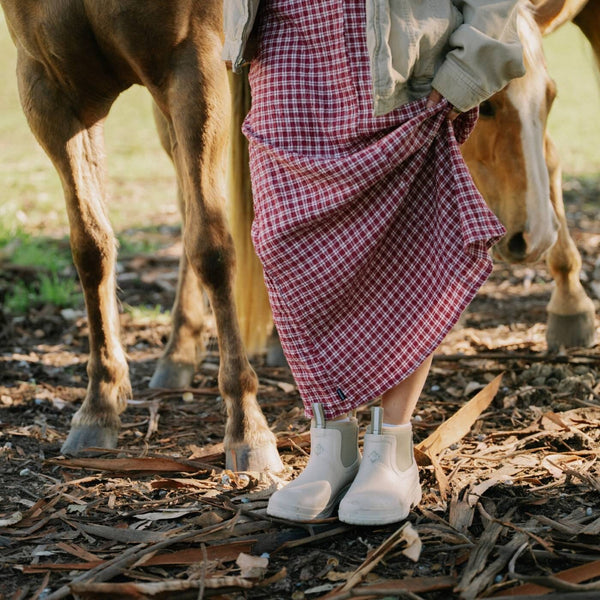 Person wearing a red plaid skirt and beige boots standing next to a horse in a natural setting.