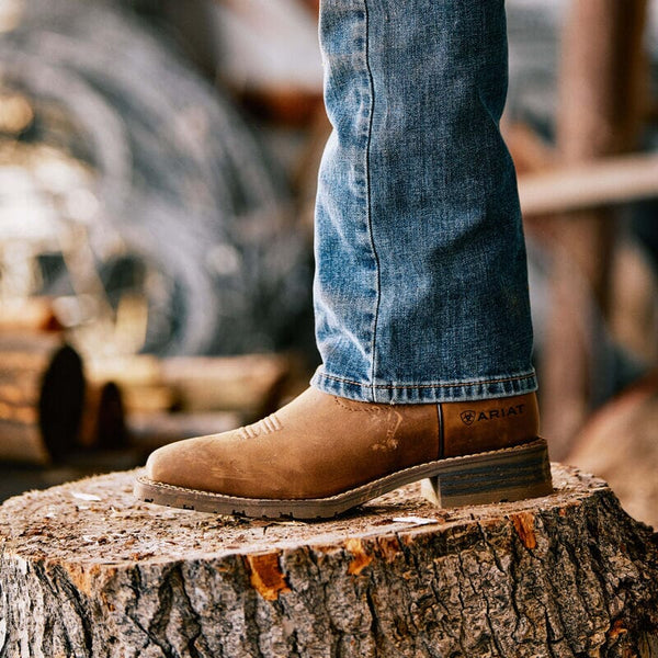 one leg propped on cut tree stump wearing light brown western boot with blue jeans over shaft