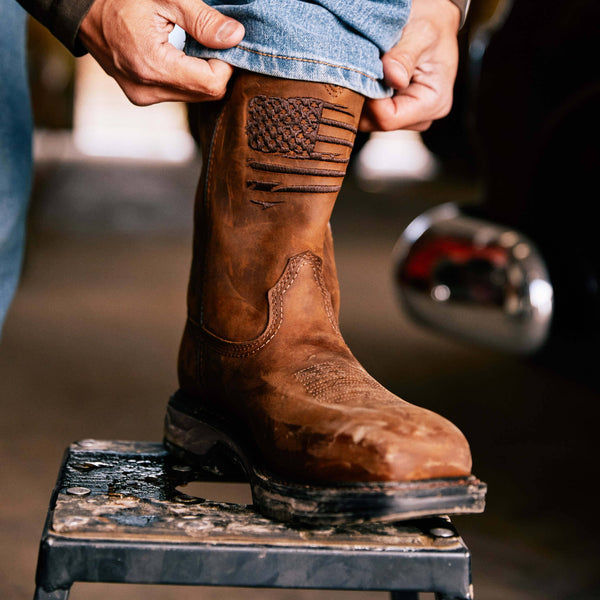 photo of hands holding bottom hem of light blue jeans over top of brown distressed western work boot with a rugged American flag embroidered across the shaft