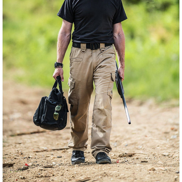 man walking with rifle and bag while wearing black boots, black shirt, and brown pants
