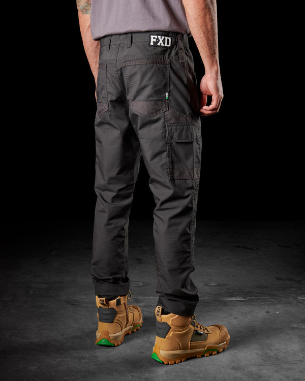 back view of man wearing black light weight work pants with FXD logo, grey tucked shirt, and tan boots