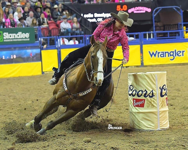 woman in western clothes and brown cowgirl hat riding brown horse around a barrel in a dirt arena