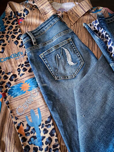 blue jeans with feather embroidered on back pocket and leopard print on hem, folded on top of leopard and desert print collared shirt.