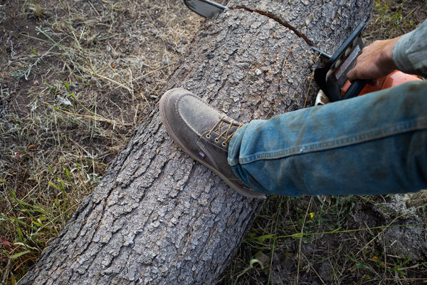 man chain sawing log with foot on log while wearing brown high top moccasin style boot with yellow and brown laces